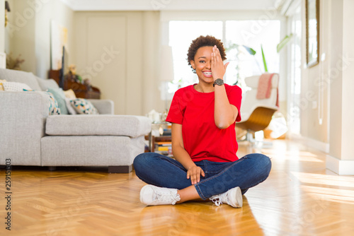 Young beautiful african american woman sitting on the floor at home covering one eye with hand with confident smile on face and surprise emotion.