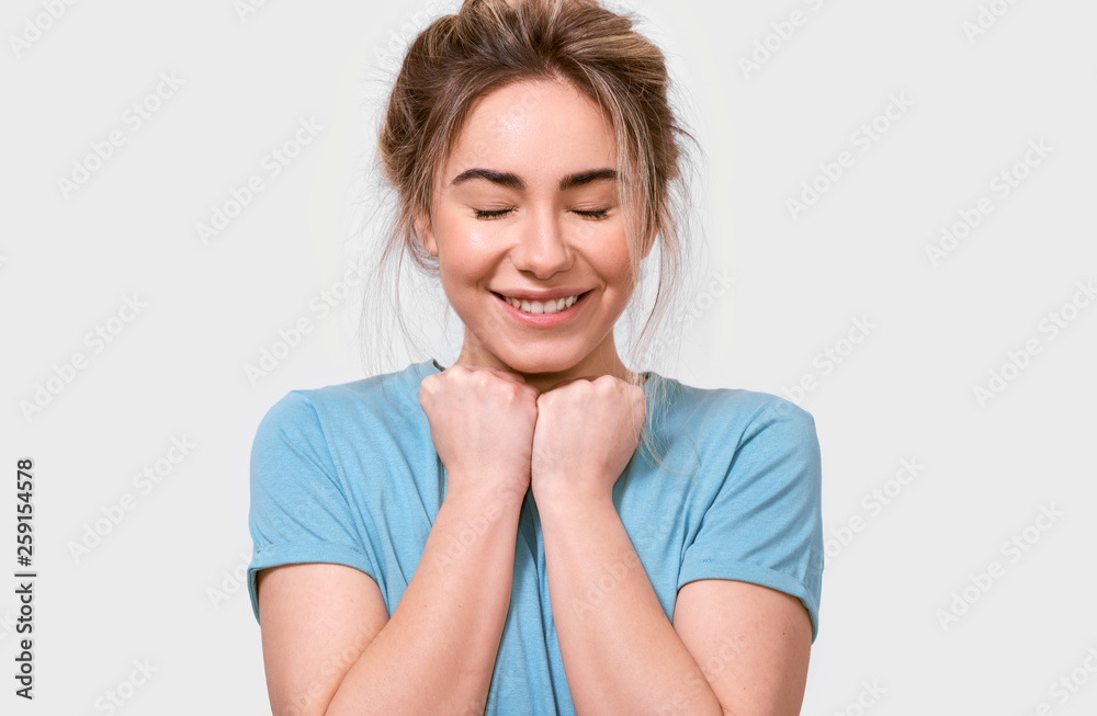 Fototapeta premium Grateful young woman smiling and keeps hands on chest with closed eyes, dressed in blue t-shirt, isolated over white background. People, emotions, feeling and body language concept