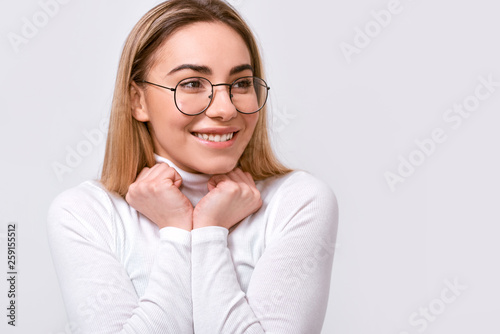 Close up portrait of adorable happy young woman smiling and keeps hands on chin, looking aside, dressed in white blouse and round eyewear, isolated over white background. People, emotions, feelings