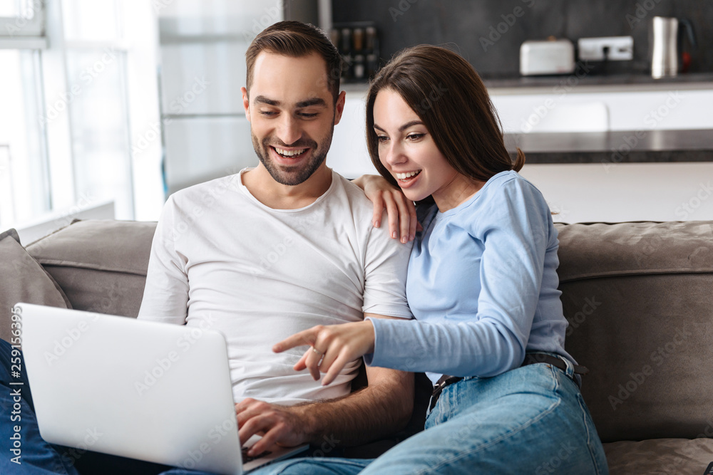 Image of happy couple using laptop together while sitting on sofa in living room at home Image of happy couple using laptop together while sitting on sofa in living room at home