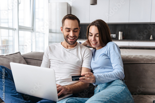 Image of lovely couple using laptop and credit card together for online shopping while sitting on sofa at home