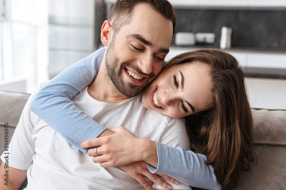 Image of couple in love hugging together at home while sitting on sofa in living room Image of couple in love hugging together at home while sitting on sofa in living room