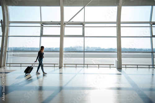 Young female pasenger in casual clothes with luggage walking near big windows in airport terminal hall. Waiting for a flight. Walking at the gate