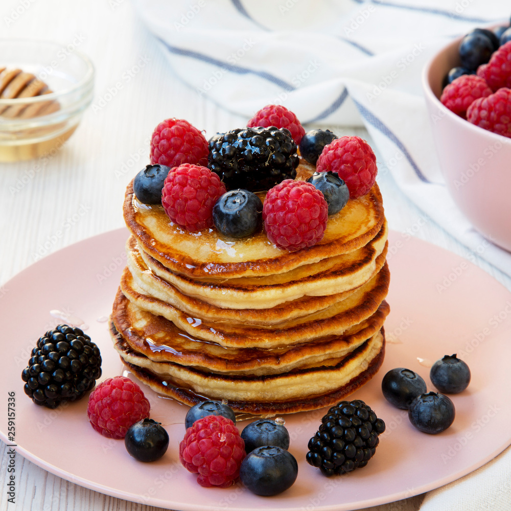 Homemade pancakes with berries and honey on a pink plate over white wooden surface, side view. Close-up. Homemade pancakes with berries and honey on a pink plate over white wooden surface, side view. Close-up.