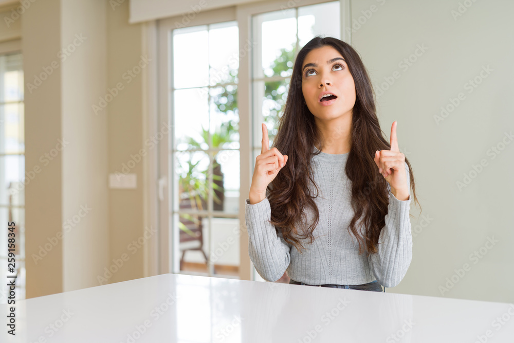 Young beautiful woman at home amazed and surprised looking up and pointing with fingers and raised arms. Young beautiful woman at home amazed and surprised looking up and pointing with fingers and raised arms.