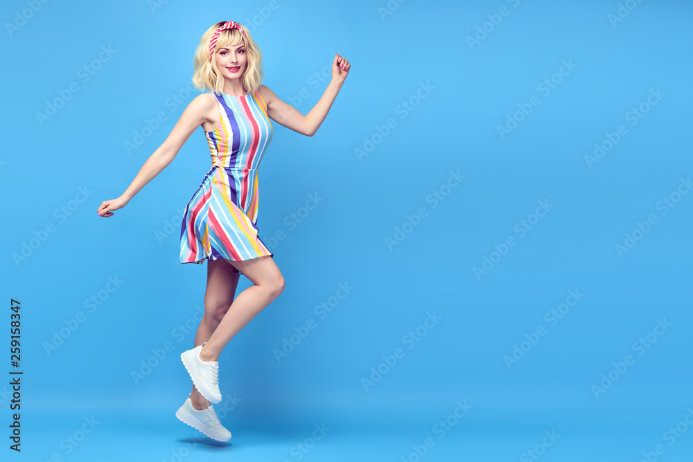 Fashionable shapely Young woman smiling on blue background. Excited blonde Girl Having Fun dance in Trendy summer dress, Stylish fashion sneakers, makeup. Happiness funny concept