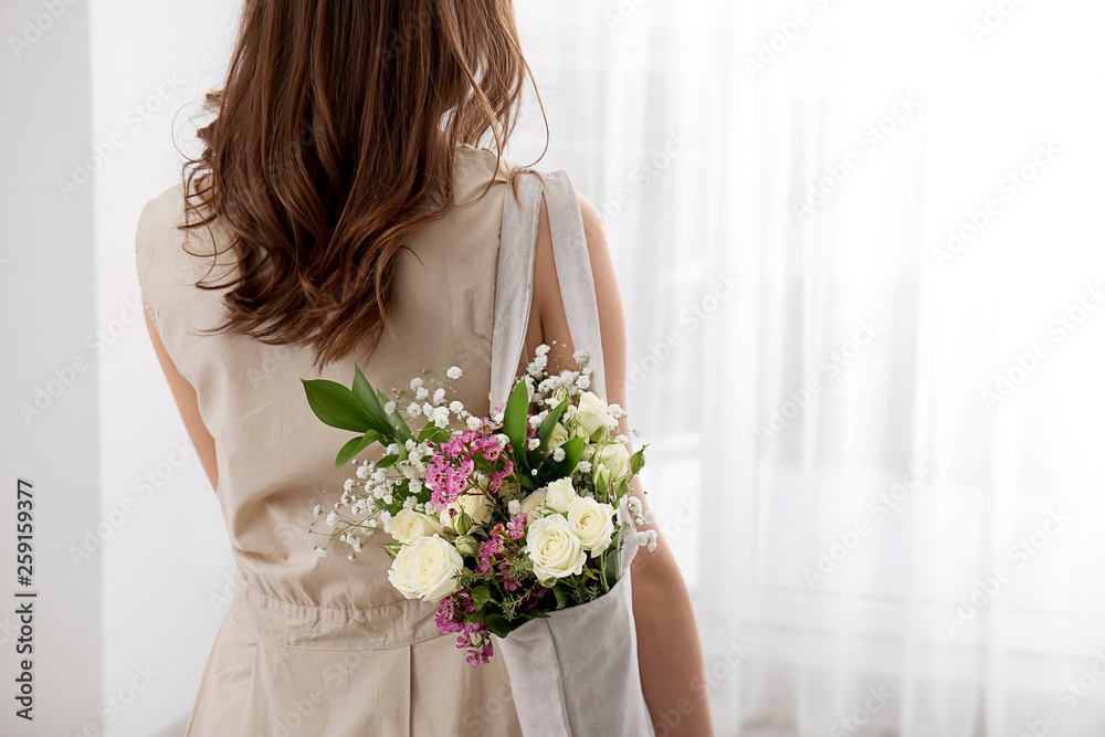 Young woman with bouquet of flowers in eco bag indoors