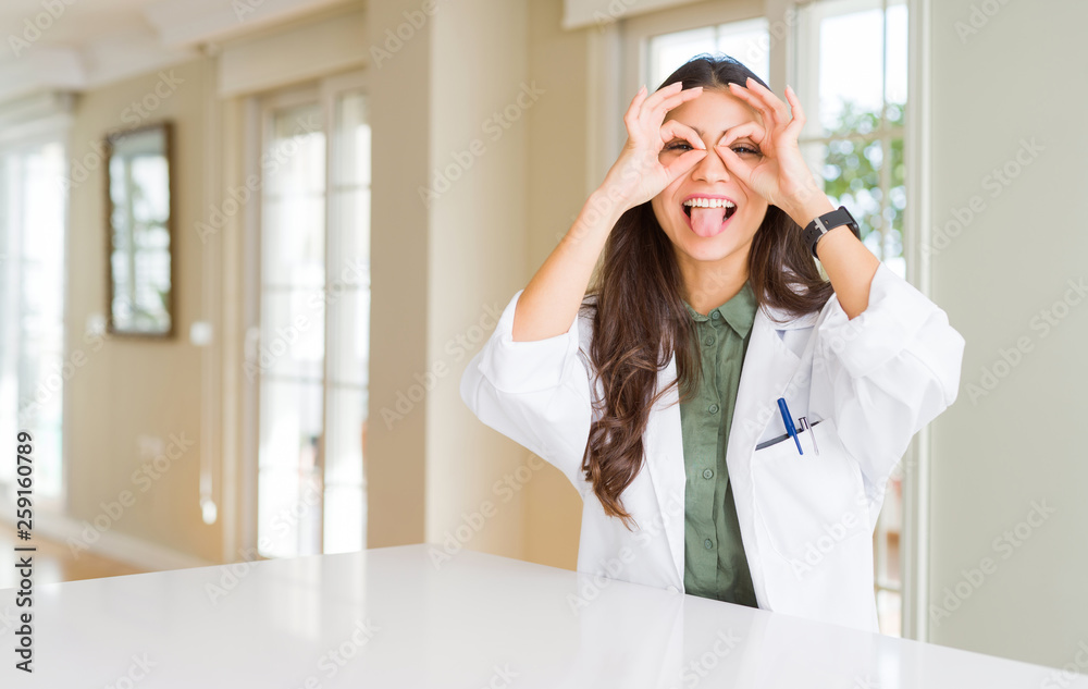 Young woman wearing medical coat at the clinic as therapist or doctor doing ok gesture like binoculars sticking tongue out, eyes looking through fingers. Crazy expression. Young woman wearing medical coat at the clinic as therapist or doctor doing ok gesture like binoculars sticking tongue out, eyes looking through fingers. Crazy expression.