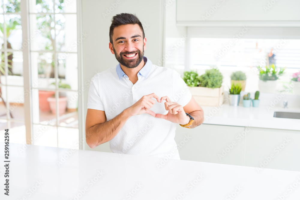 Handsome hispanic man casual white t-shirt at home smiling in love showing heart symbol and shape with hands. Romantic concept.