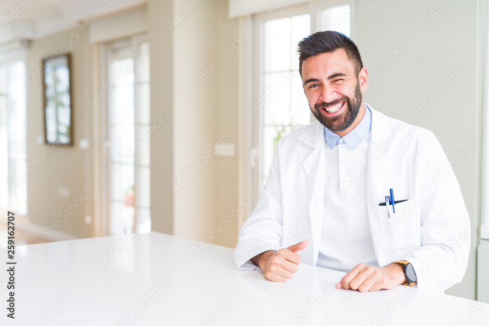 Handsome hispanic doctor or therapist man wearing medical coat at the clinic winking looking at the camera with sexy expression, cheerful and happy face.