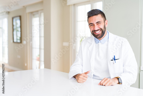 Handsome hispanic doctor or therapist man wearing medical coat at the clinic winking looking at the camera with sexy expression  cheerful and happy face.