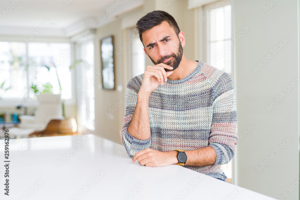 Handsome hispanic man wearing casual sweater at home with hand on chin thinking about question, pensive expression. Smiling with thoughtful face. Doubt concept.