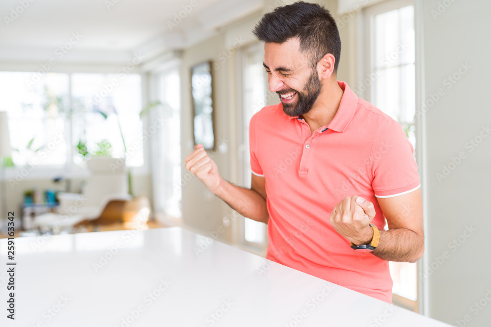 Handsome hispanic man wearing casual t-shirt at home very happy and excited doing winner gesture with arms raised, smiling and screaming for success. Celebration concept. Handsome hispanic man wearing casual t-shirt at home very happy and excited doing winner gesture with arms raised, smiling and screaming for success. Celebration concept.