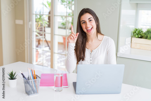 Beautiful young student woman studying for university using laptop and notebook surprised with an idea or question pointing finger with happy face, number one