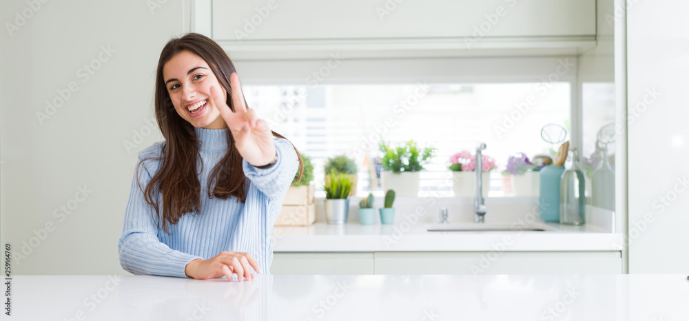 Wide angle picture of beautiful young woman sitting on white table at home smiling looking to the camera showing fingers doing victory sign. Number two. Wide angle picture of beautiful young woman sitting on white table at home smiling looking to the camera showing fingers doing victory sign. Number two.