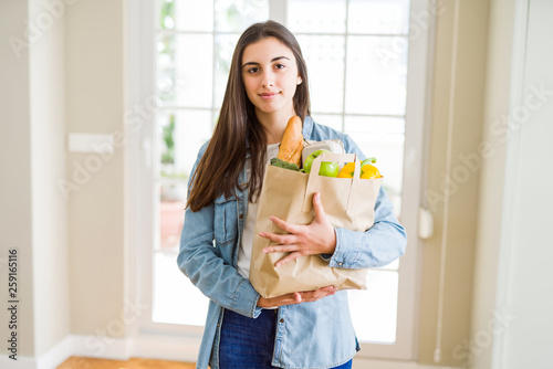 Beautiful young woman holding paper bag full of healthy groceries with a confident expression on smart face thinking serious