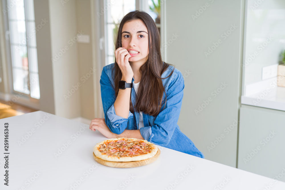 Beautiful young woman eating homemade tasty pizza at the kitchen looking stressed and nervous with hands on mouth biting nails. Anxiety problem. Beautiful young woman eating homemade tasty pizza at the kitchen looking stressed and nervous with hands on mouth biting nails. Anxiety problem.