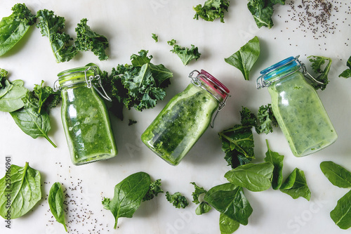 Variaty of green spinach kale apple smoothies in glass bottles with ingredients above over white marble background. Healthy organic eating. Flat lay, space photo