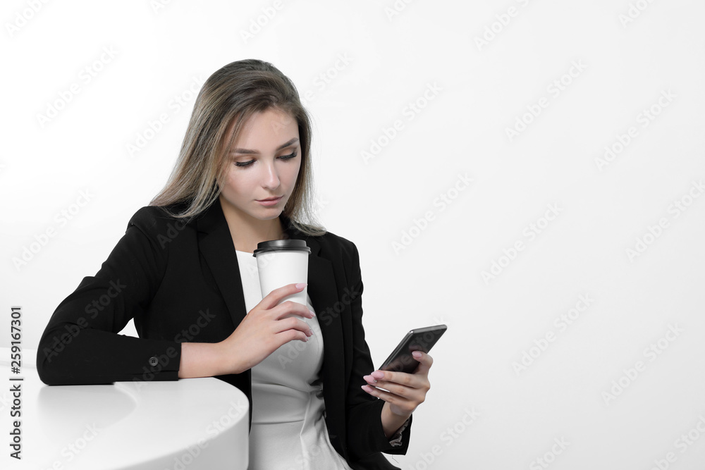 A young beautiful woman in a black suit and white blouse is sitting at the office table. Drinks a hot drink of coffee from a white paper cup. Writes a message on a smartphone, communicates and smiles. A young beautiful woman in a black suit and white blouse is sitting at the office table. Drinks a hot drink of coffee from a white paper cup. Writes a message on a smartphone, communicates and smiles.