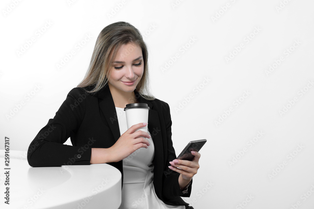 A young beautiful woman in a black suit and white blouse is sitting at the office table. Drinks a hot drink of coffee from a white paper cup. Writes a message on a smartphone, communicates and smiles. A young beautiful woman in a black suit and white blouse is sitting at the office table. Drinks a hot drink of coffee from a white paper cup. Writes a message on a smartphone, communicates and smiles.