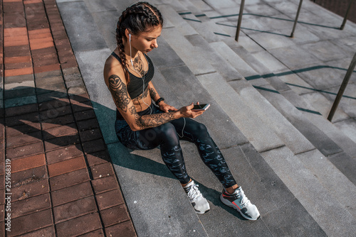 Athlete girl tattoos, sitting on steps summer city, hand smartphone, listening music with headphones. Resting after jogging workout. Concept of motivation, active life position, healthy lifestyle.