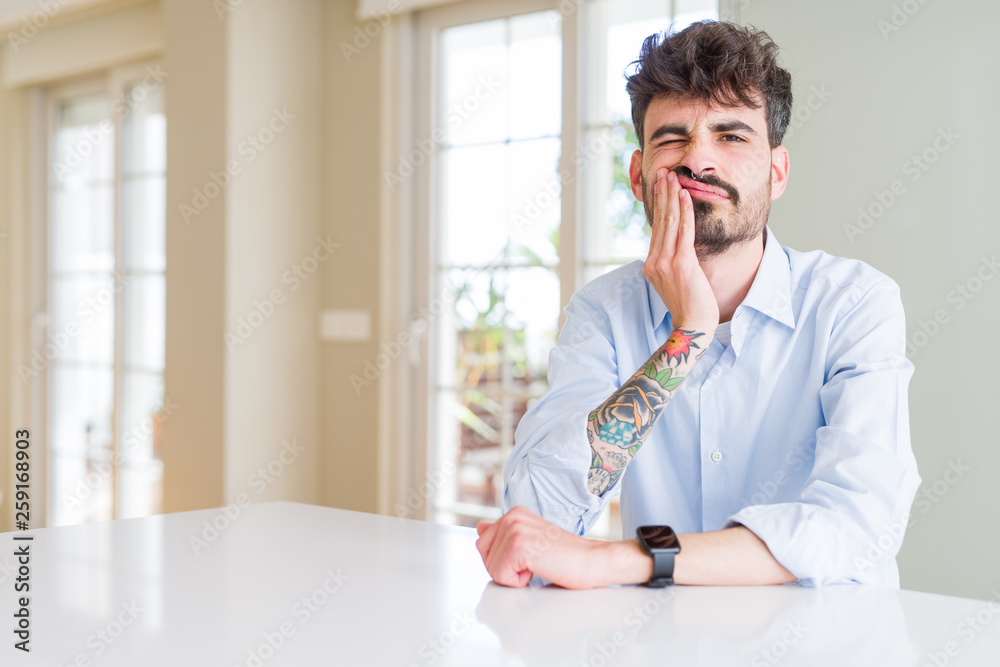 Young businesss man sitting on white table touching mouth with hand with painful expression because of toothache or dental illness on teeth. Dentist concept.