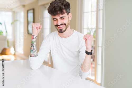 Young man wearing casual shirt sitting on white table very happy and excited doing winner gesture with arms raised, smiling and screaming for success. Celebration concept.