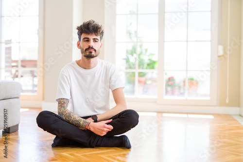 Young man working sitting casual on the floor at home with serious expression on face. Simple and natural looking at the camera.