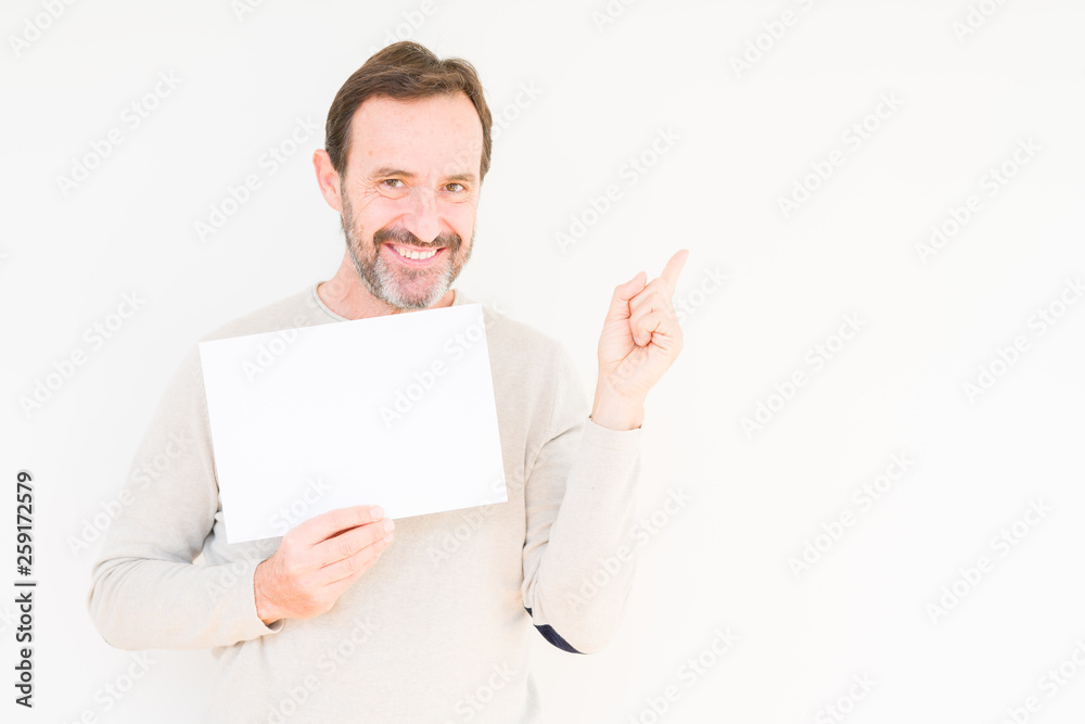 Senior man holding blank paper sheet over isolated background very happy pointing with hand and finger to the side Senior man holding blank paper sheet over isolated background very happy pointing with hand and finger to the side