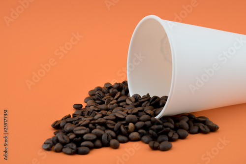 Paper coffee cup with spilled coffee beans, orange background. Coffee beans.