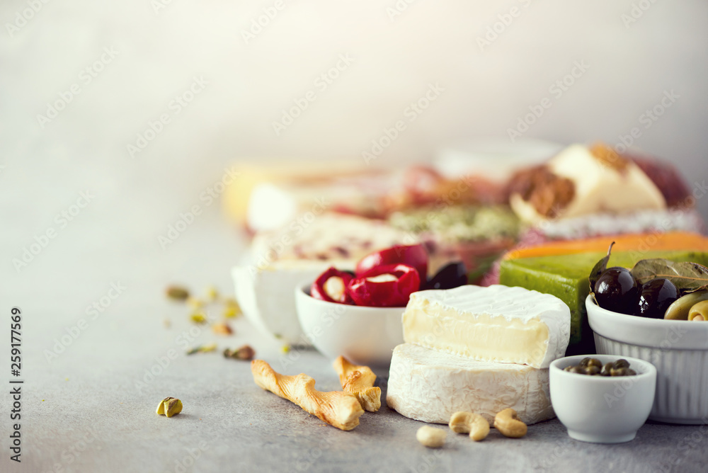 Assortment of hard, semi-soft and soft cheeses with olives, grissini bread sticks, capers, grape, on grey concrete backgound. Top view, copy space, flat lay. Cheese selection appetizer plate. Assortment of hard, semi-soft and soft cheeses with olives, grissini bread sticks, capers, grape, on grey concrete backgound. Top view, copy space, flat lay. Cheese selection appetizer plate.