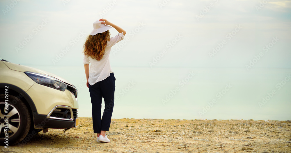 Fototapeta premium Happy stylish young woman traveler on beach road near white crossover car, holding hat in hand. Banner. Travel, summer vacation, holiday, freedom concept. Digital detox