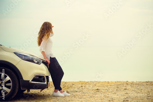Happy stylish young woman traveler on beach road sitting on white crossover car, holding hat in hand. Banner. Travel, summer vacation, holiday, freedom concept. Digital detox © jchizhe