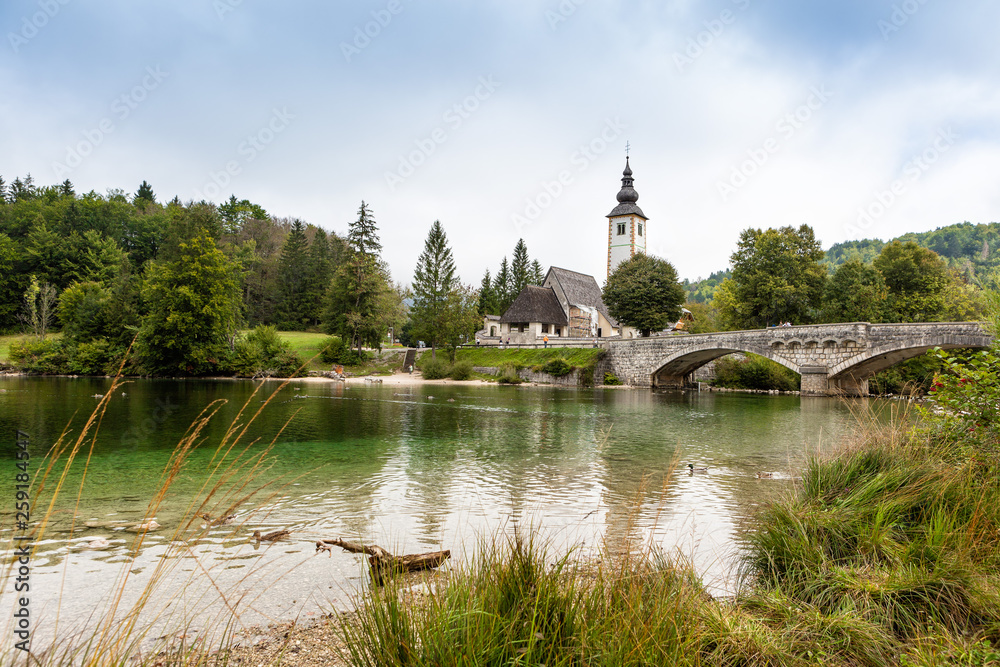 Lake Bohinj in Slovenia on an autumn morning. The view from the coast overgrown with grass to the bridge, forest and church Lake Bohinj in Slovenia on an autumn morning. The view from the coast overgrown with grass to the bridge, forest and church