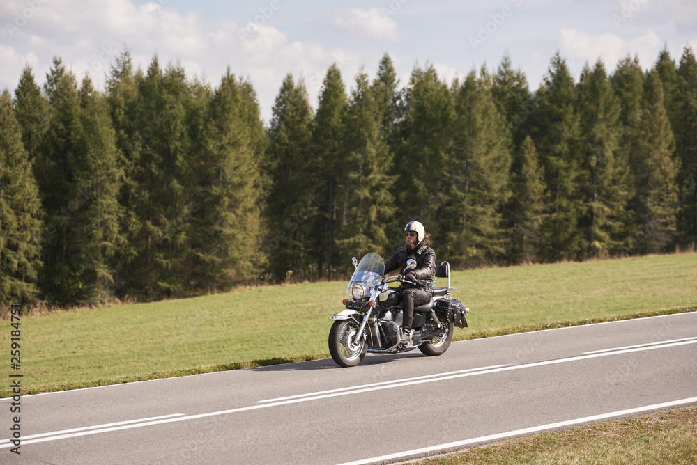 Bearded motorcyclist in helmet, sunglasses and black leather clothing riding cruiser motorbike down empty asphalt road on sunny summer day on background of tall dense spruce trees forest.