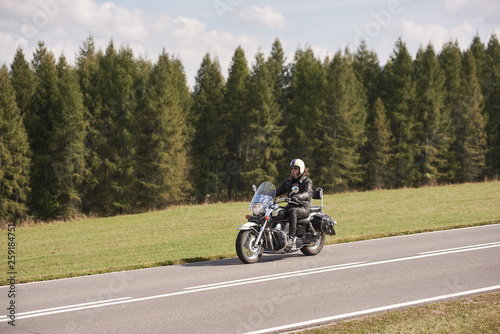 Bearded motorcyclist in helmet  sunglasses and black leather clothing riding cruiser motorbike down empty asphalt road on sunny summer day on background of tall dense spruce trees forest.