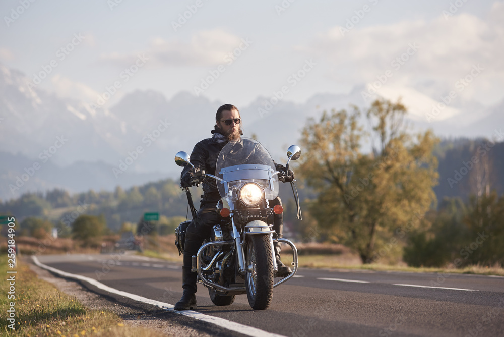 Portrait of handsome bearded biker in black leather jacket on cruiser motorcycle on country roadside on blurred background of green woody hills, distant white mountain peaks.