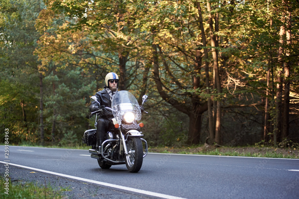 Handsome bearded biker in sunglasses, helmet and black leather clothing riding cruiser motorcycle along asphalt road winding among tall green trees on sunny summer evening.