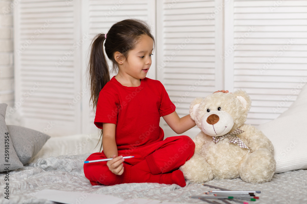 cute little child girl in red pajamas with teddy bear draws with pencils at home cute little child girl in red pajamas with teddy bear draws with pencils at home