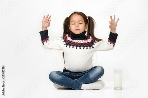 Portrait of cute little child girl sitting in lotus position and a glass of fresh milk is standing nearby