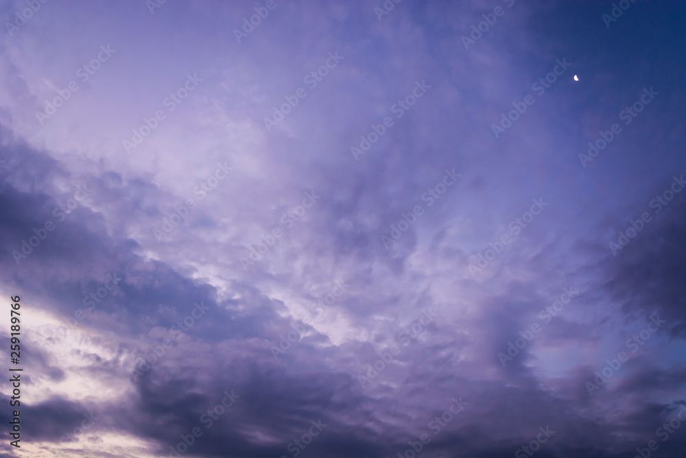 Fototapeta premium Dramatic evening sky with purple and blue dark clouds like Cirrocumulus, Altocumulus and Cumulus. Small crescent moon behind cloudscape. Wind and motion. 