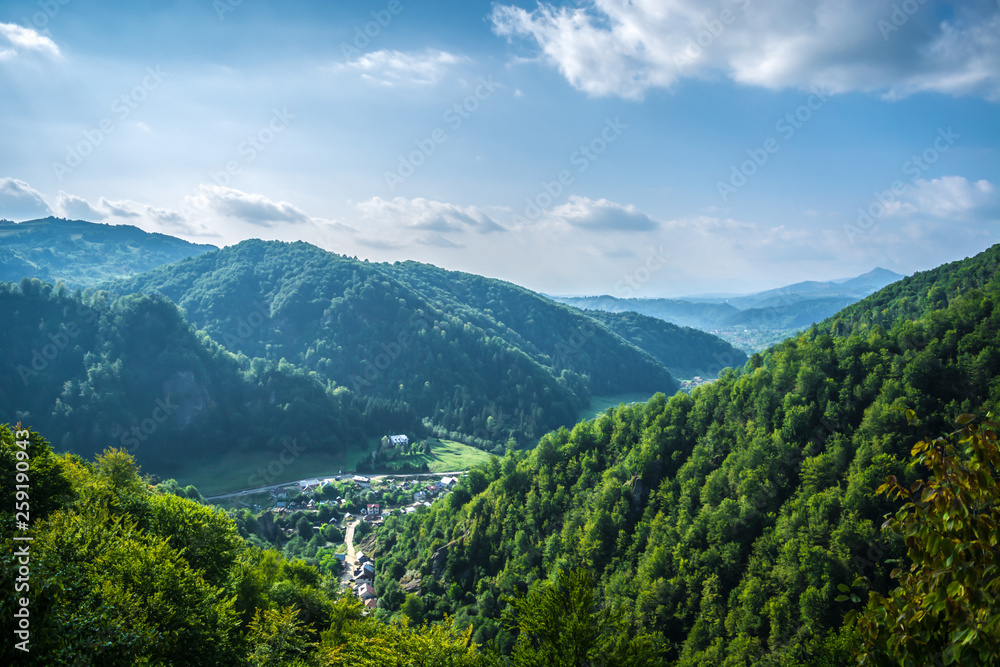 Village surrounded by forested mountains with blue sky in Romania, Eastern Europe. Concept of travel, escape society, nature relaxing, remote places, traditional landscape. Carpathian Mountains.