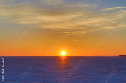 The sunset or sunrise. The cloudy sky cloured in red  orange  crimson  purple  violet and blue bright and vivid coloures with setting or rising sun over the sea  lake or bay covered with ice and snow