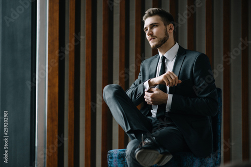 Young handsome man posing for a fashion shoot in a studio. Fashion as a lifestyle. Man wearing a beard. Model sitting on a sofa in tuxedo. Successful fashionable businessman. Business look advertising