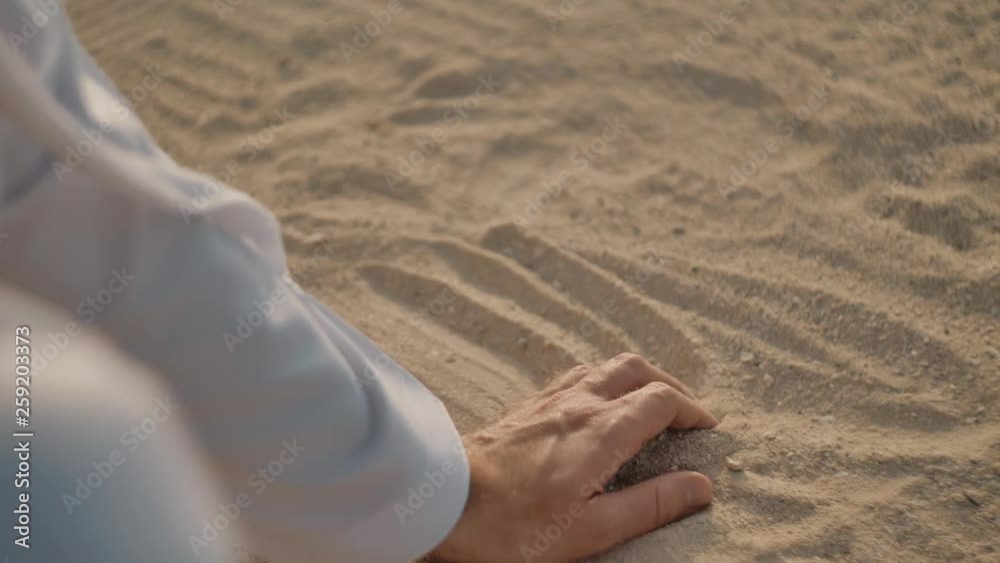 Caucasian man in white clothes, runs his hand over the sand, takes a handful of it into a fist, then opens his hand and sand pours through his fingers. The sun is shining.