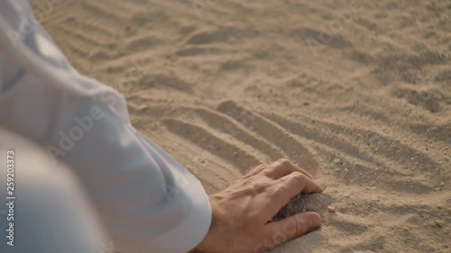 Caucasian man in white clothes, runs his hand over the sand, takes a handful of it into a fist, then opens his hand and sand pours through his fingers. The sun is shining.