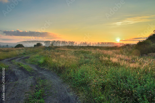 morning, rising sun over the field and dirt road, with trees and country buildings