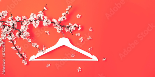 White wooden hanger hanging on the spring flowering branch on coral background. Spring sale concept discount store shopping empty hanger. Creative fashion beauty banner. Flat lay top view copy space.