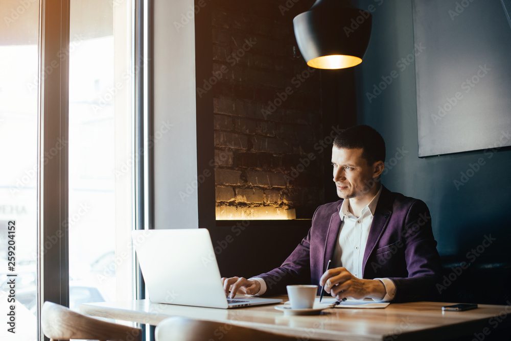 Young handsome man sitting in office with cup of coffee and working on project connected with modern cyber technologies. Businessman with notebook trying to keep deadline in digital marketing sphere. Young handsome man sitting in office with cup of coffee and working on project connected with modern cyber technologies. Businessman with notebook trying to keep deadline in digital marketing sphere.