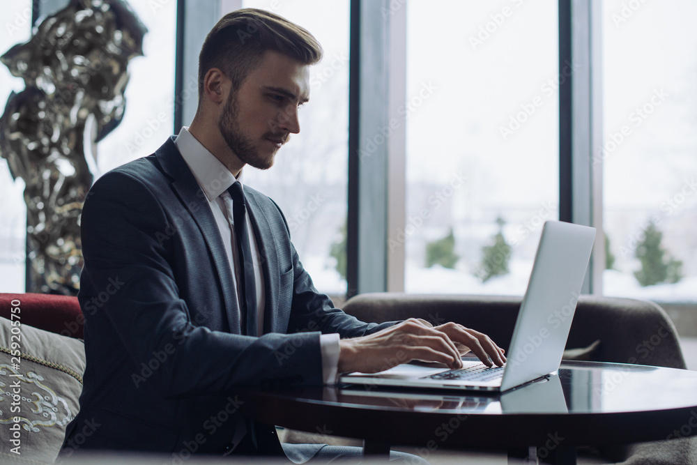 Young handsome man sitting in office with cup of coffee and working on project connected with modern cyber technologies. Businessman with notebook trying to keep deadline in digital marketing sphere.